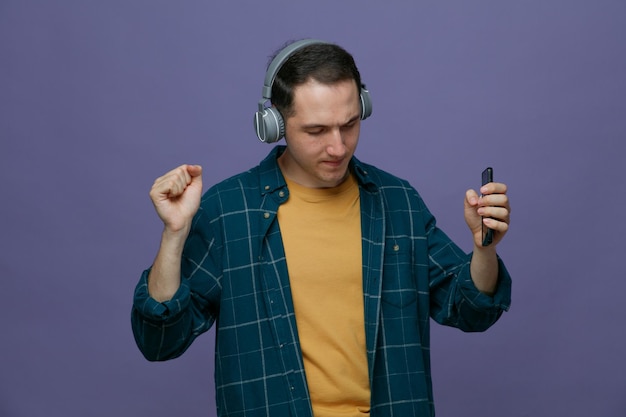 Pleased young male student wearing headphones holding mobile phone listening to music dancing with closed eyes isolated on purple background