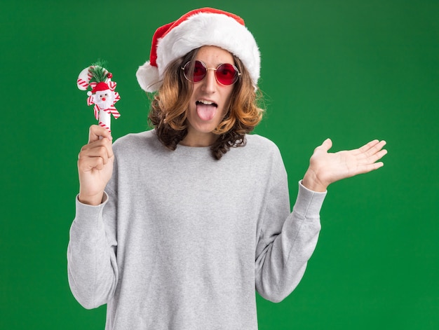 Free photo pleased young man wearing christmas santa hat and red glasses holding christmas candy cane happy and cheerful sticking out tongue presenting with arm of hand standing over green background