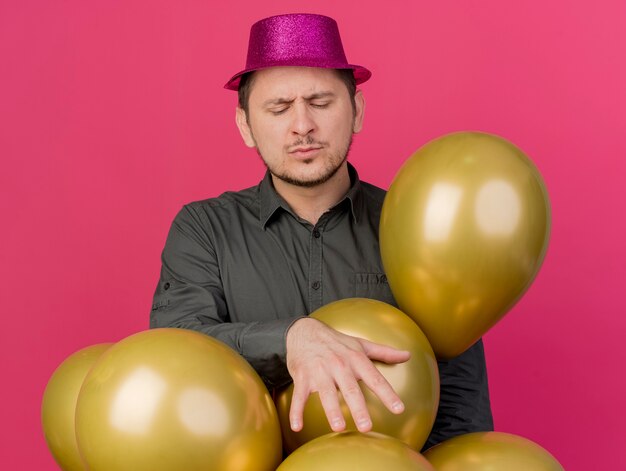 Pleased young party guy with closed eyes wearing pink hat standing among balloons isolated on pink