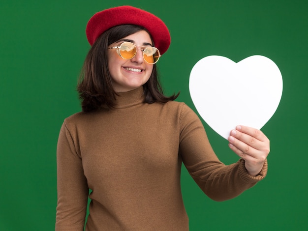 Pleased young pretty caucasian girl with beret hat in sun glasses holding heart shape on green