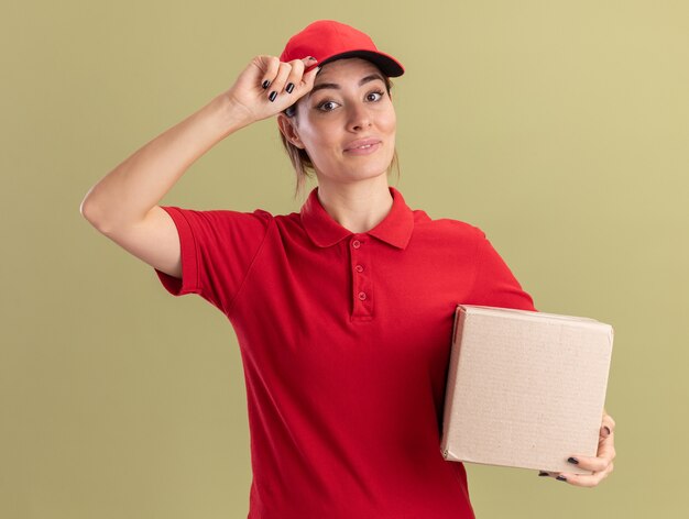 Pleased young pretty delivery girl in uniform puts hand on cap and holds cardbox on olive green