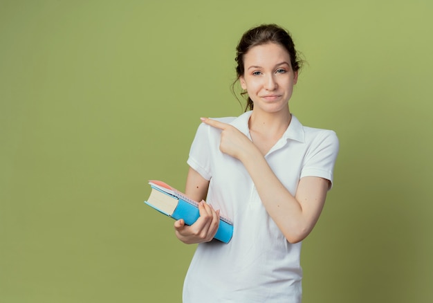 Pleased young pretty female student holding book and note pad pointing at side isolated on olive green background with copy space