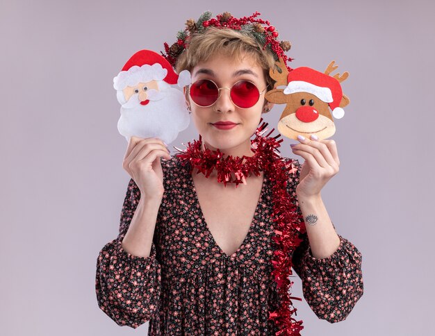 pleased young pretty girl wearing christmas head wreath and tinsel garland around neck with glasses holding christmas reindeer and santa claus paper ornaments near head looking at side isolated on white wall