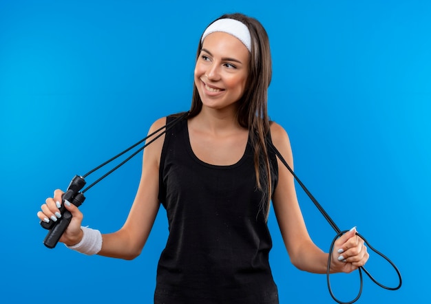 Pleased young pretty sporty girl wearing headband and wristband holding jumping rope around her neck looking at side isolated on blue space