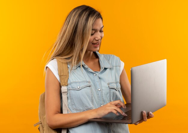 Pleased young pretty student girl wearing back bag using laptop isolated on orange wall