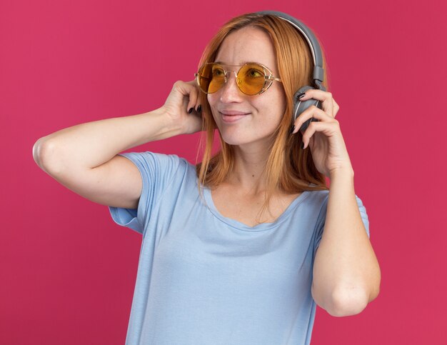 Pleased young redhead ginger girl with freckles in sun glasses and on headphones looking at side on pink