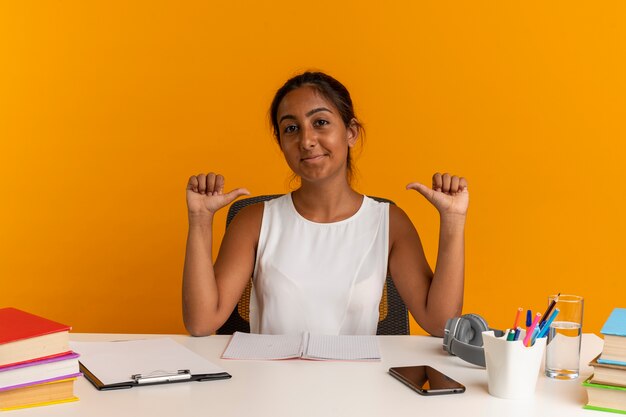 Pleased young schoolgirl sitting at desk with school tools points to herself isolated on orange wall