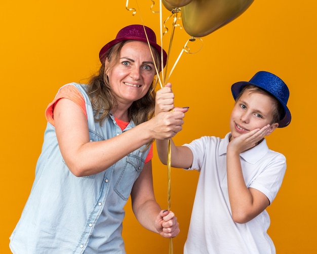 pleased young slavic boy with blue party hat putting hand on face and holding helium balloons with his mother wearing purple party hat isolated on orange wall with copy space