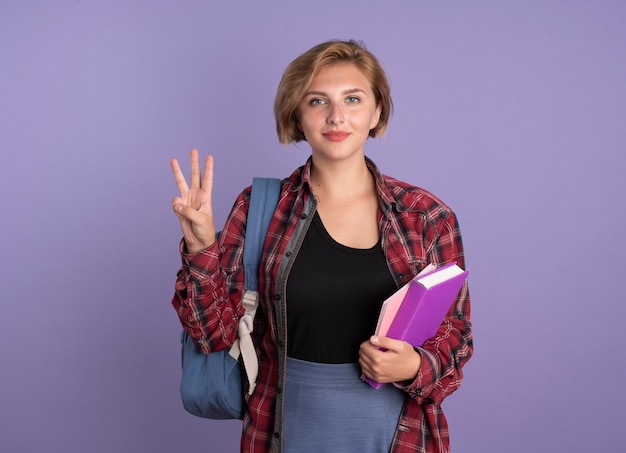 Pleased young slavic student girl wearing backpack holds book and notebook gestures three with fingers 