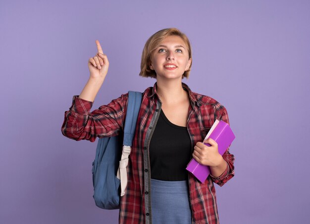 Pleased young slavic student girl wearing backpack holds book and notebook points up 