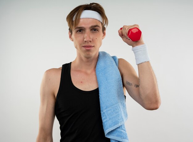 Pleased young sporty guy wearing headband and wristband exercising with dumbbell with towel on shoulder isolated on white wall