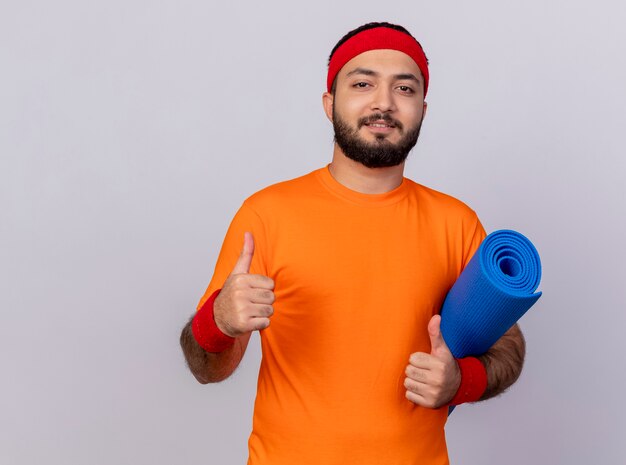 Pleased young sporty man wearing headband and wristband holding yoga mat