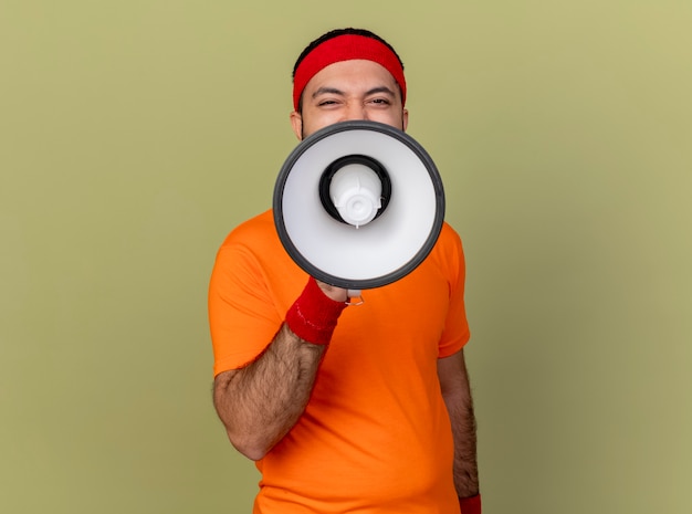 Free photo pleased young sporty man wearing headband and wristband speaks on loudspeaker isolated on olive green