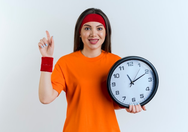 Pleased young sporty woman wearing headband and wristbands holding clock looking pointing up 