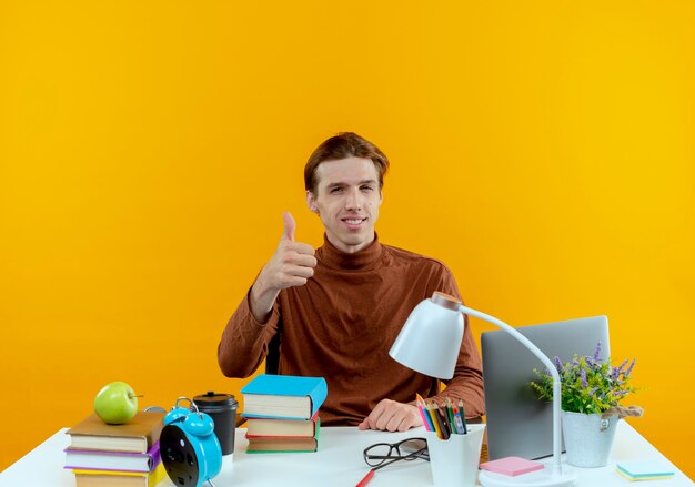 Pleased young student boy sitting at desk with school tools his thumb up isolated on yellow wall