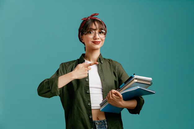 Pleased young student girl wearing bandana glasses holding large note pads with pen looking at camera pointing at note pads isolated on blue background