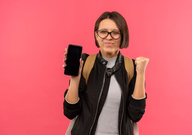 Pleased young student girl wearing glasses and back bag holding mobile phone clenching fist with closed eyes isolated on pink wall