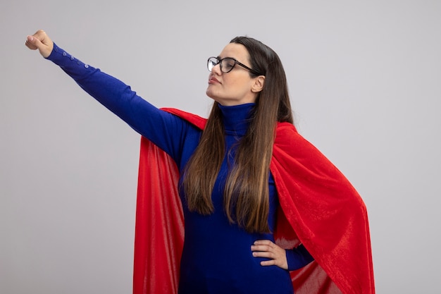 Pleased young superhero girl looking at side wearing glasses raising hand isolated on white background