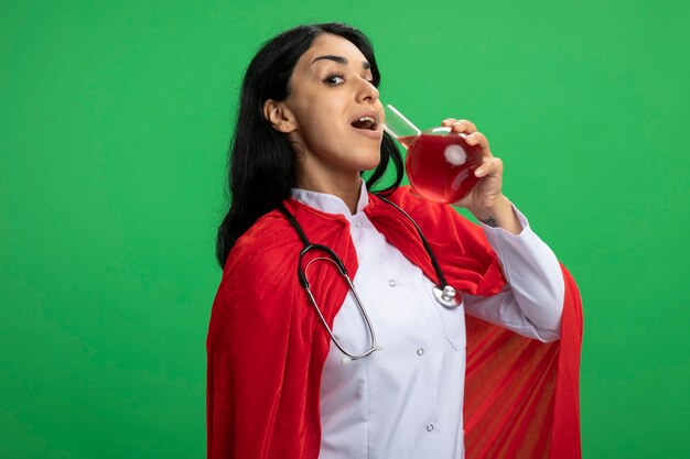 Pleased young superhero girl wearing medical robe with stethoscope holding and drinks chemistry glass bottle filled with red liquid isolated on green