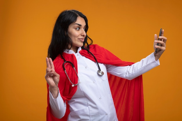 Pleased young superhero girl wearing medical robe with stethoscope take a selfie and showing peace gesture isolated on orange wall