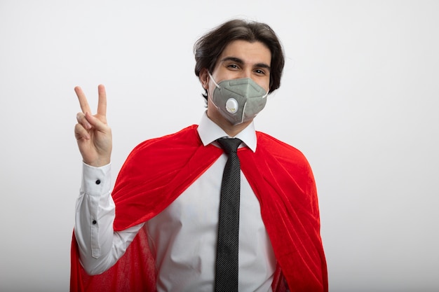 Free photo pleased young superhero guy looking at camera wearing tie and medical mask showing peace gesture isolated on white background