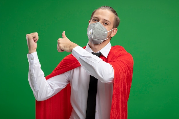 Free photo pleased young superhero guy wearing medical mask points at back isolated on green