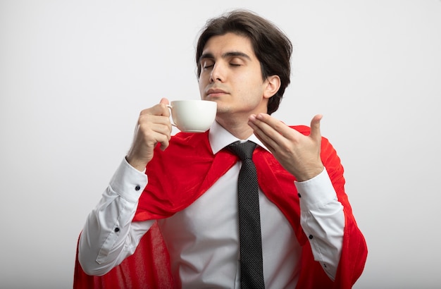 Pleased young superhero guy with closed eyes wearing tie sniffing coffee isolated on white