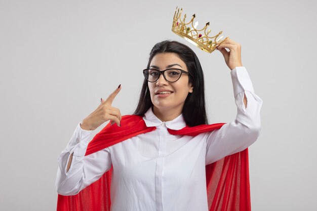 Pleased young superwoman wearing glasses holding crown above head looking at front pointing at crown isolated on white wall