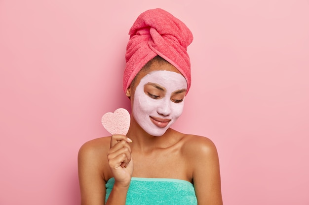 Pleased young woman focused down, applies facial clay mask, holds cosmetic sponge for removing makeup, shows bare shoulders, wrapped in bath towel, isolated on pink studio wall