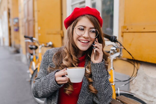 Pleased young woman in gray jacket talking on phone and drinking coffee in street cafe