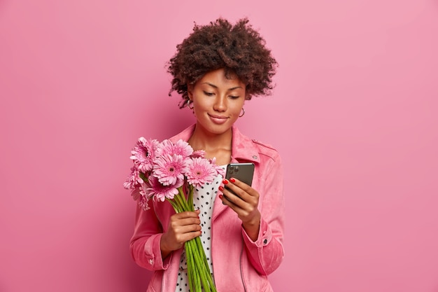 Free photo pleased young woman has afro hair, got gerbera bouquet as present, poses with beautiful flowers and smartphone in hands, sends messages online, gets surprise gift