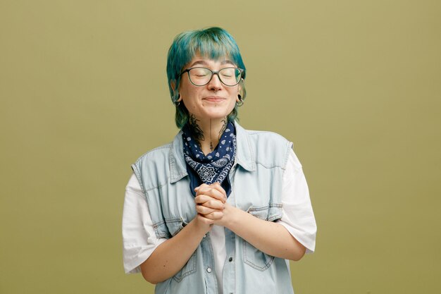 Pleased young woman wearing glasses bandana on neck keeping hands together with closed eyes isolated on olive green background