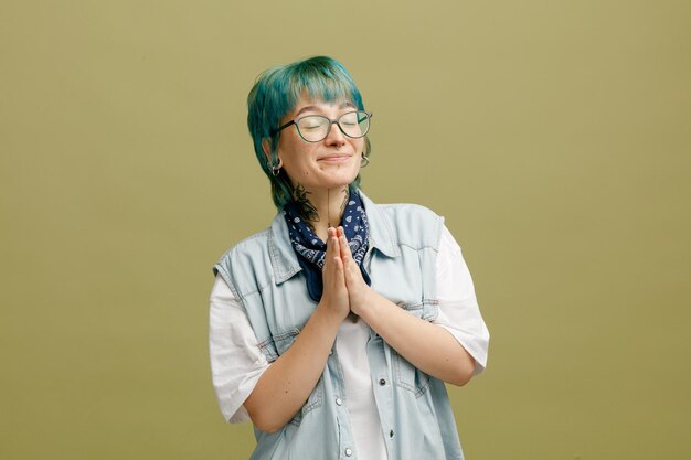 Pleased young woman wearing glasses bandana on neck showing namaste gesture with closed eyes isolated on olive green background