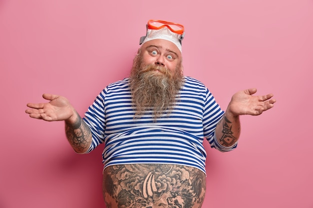 Plump hesitant bearded adult man spreads hands with doubt, wears snorkeling equipment, has thick beard, dressed in sailor undresized shirt