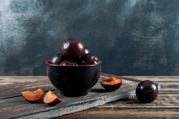 Plums with slices in a bowl with wooden board side view on a dark wooden and grey grunge background