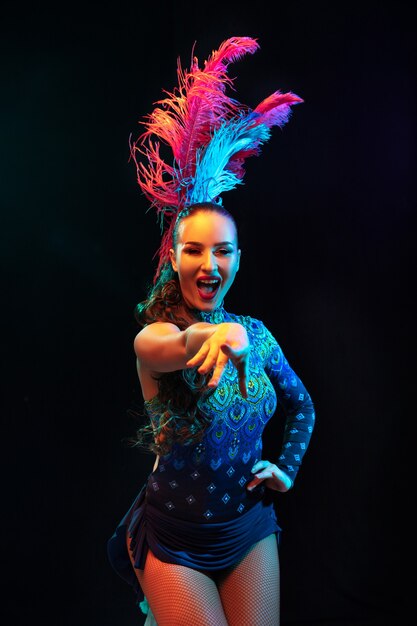 Pointing. Beautiful young woman in carnival, stylish masquerade costume with feathers on black wall in neon light. Copyspace for ad. Holidays celebration, dancing, fashion. Festive time, party.