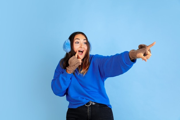 Pointing. Caucasian woman's portrait on blue studio background. Beautiful female model in warm clothes. Concept of human emotions, facial expression, sales, ad. Winter mood, Christmas time, holidays.