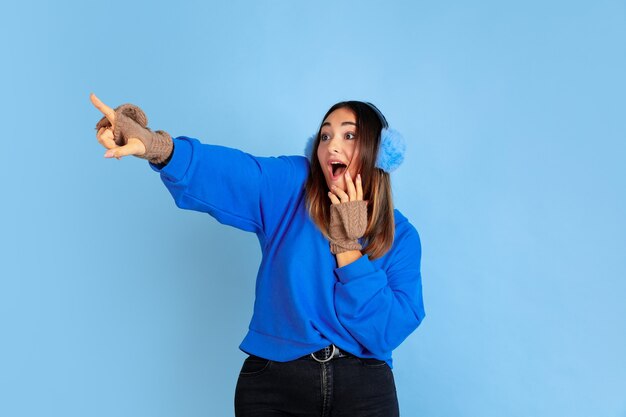 Pointing. Caucasian woman's portrait on blue studio background. Beautiful female model in warm clothes. Concept of human emotions, facial expression, sales, ad. Winter mood, Christmas time, holidays.