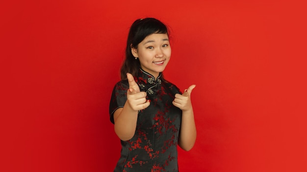 Free photo pointing on, choosing, smiling. happy chinese new year 2020. asian young girl's portrait on red background. female model in traditional clothes looks happy. celebration, human emotions. copyspace.