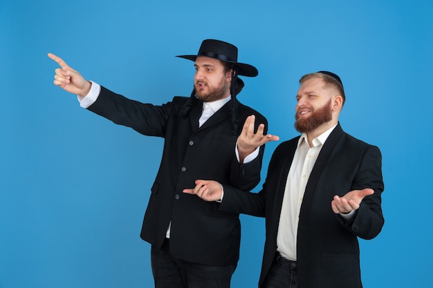 Free Photo pointing, inviting. portrait of a young orthodox jewish men isolated on blue wall. purim, business, festival, holiday, celebration pesach or passover, judaism, religion concept.