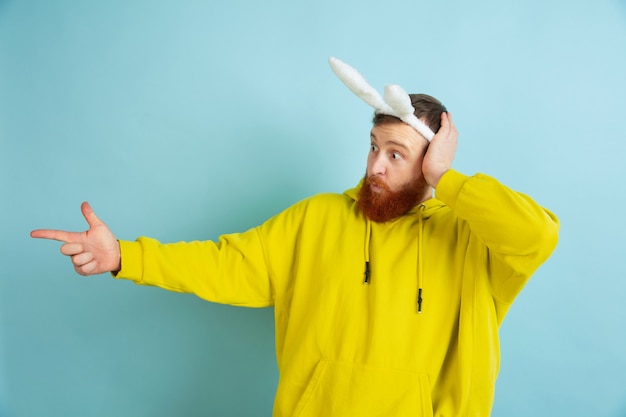 Free Photo pointing at side. caucasian man as an easter bunny with bright casual clothes on blue studio background.