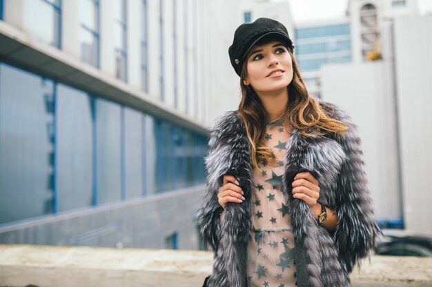 Portrair of fashionable woman walking in city in warm fur coat wearing black cap