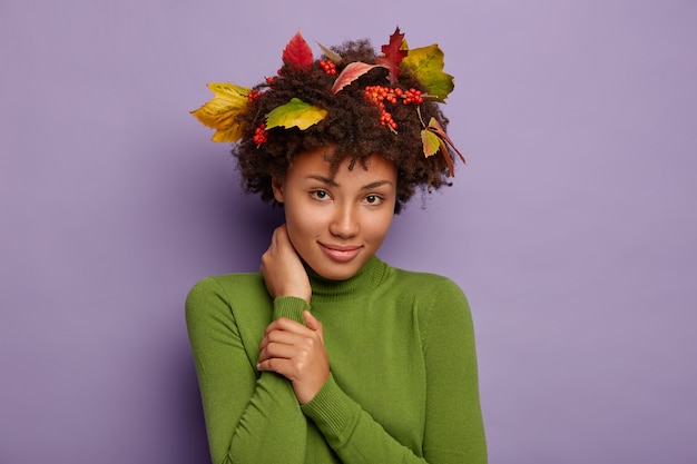 Free photo portrait of adorable afro american lady touches neck, looks calmly at camera, wears casual poloneck, autumn foliage, berries in curly hair for perfect hairstyle satisfied