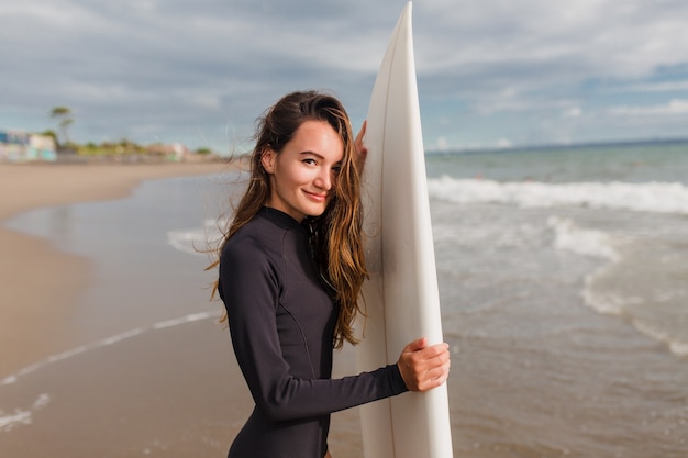 Portrait of adorable friendly young woman with long light-brown hair and big eyes stands on shore of the ocean with happy true emotions and preparing for surf lesson