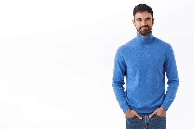 Portrait of adult handsome man with beard, hold hands in jeans pockets while smiling with friendly carefree expression, standing white wall casual normal pose