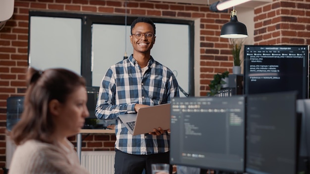 Free photo portrait of african american software developer holding laptop looking up and smiling at camera. programer typing on portable computer next to coworker coding cyber security agency.