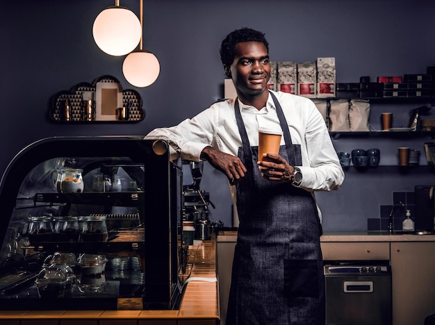 Free Photo portrait of an african barista holding a cup with coffee while leaning on a counter in a coffee shop