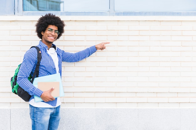 Portrait of an african smiling male student holding books in hand pointing the finger on white painted wall