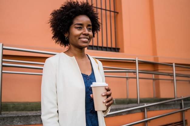 Portrait of afro business woman holding a cup of coffee while standing outdoors on the street. Business and urban concept.