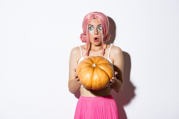 Portrait of amazed young party girl in pink wig, holding big pumpkin and looking left in awe, celebrating halloween, standing.